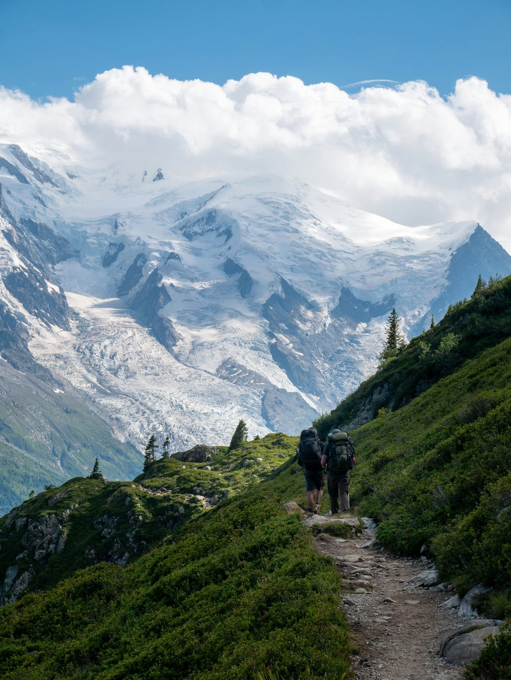 Two hikers walking a mountain trail with snow-capped peaks behind them