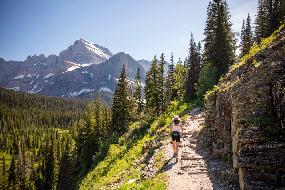 Woman hiking a mountain trail on a sunny day with snow-capped peaks behind her